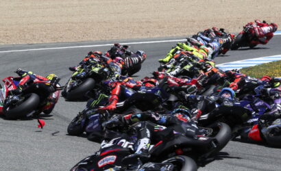 Group of racing motorcycles leaning into a tight corner on a track, riders in colorful gear close together.