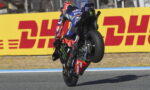 MotoGP rider in blue Yamaha livery performing a wheelie on a race track, sponsor logos visible, DHL barrier in the background.