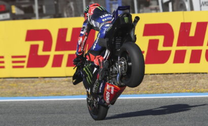 MotoGP rider in blue Yamaha livery performing a wheelie on a race track, sponsor logos visible, DHL barrier in the background.