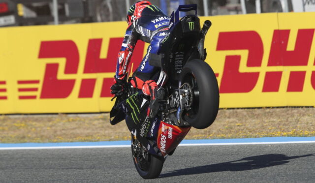 MotoGP rider in blue Yamaha livery performing a wheelie on a race track, sponsor logos visible, DHL barrier in the background.