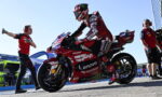 MotoGP rider in red Ducati livery sits on a racing bike with Lenovo sponsorship, surrounded by pit crew on a sunny track.