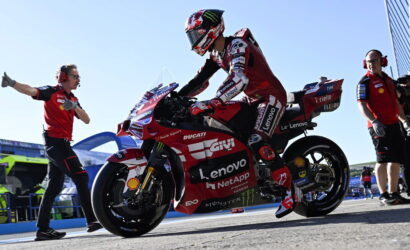 MotoGP rider in red Ducati livery sits on a racing bike with Lenovo sponsorship, surrounded by pit crew on a sunny track.