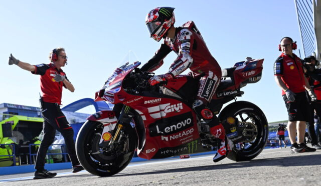MotoGP rider in red Ducati livery sits on a racing bike with Lenovo sponsorship, surrounded by pit crew on a sunny track.
