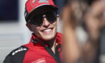 Smiling man in a red racing cap and sunglasses, with Lenovo and Autogrill logos visible on his outfit.