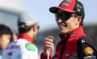 Racing driver in red Lenovo-sponsored jacket and cap, wearing dark sunglasses, smiling while holding a microphone at the track.