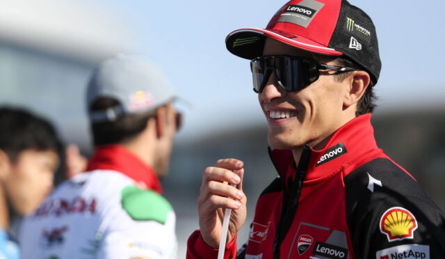 Racing driver in red Lenovo-sponsored jacket and cap, wearing dark sunglasses, smiling while holding a microphone at the track.