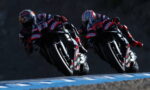Two motorcycle racers lean into a bend on a race track, wearing matching red and black racing suits.