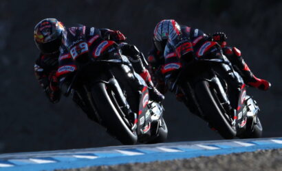 Two motorcycle racers lean into a bend on a race track, wearing matching red and black racing suits.