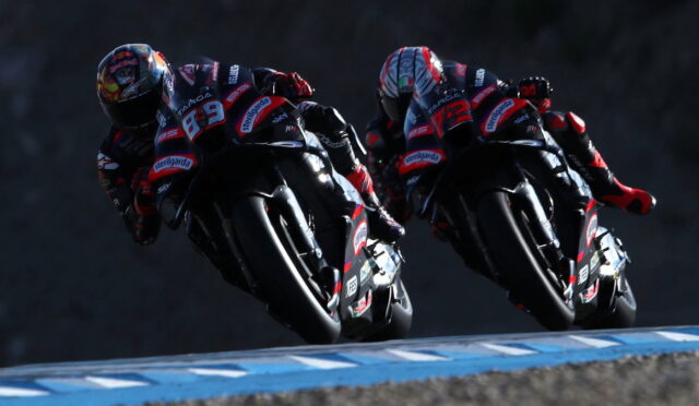 Two motorcycle racers lean into a bend on a race track, wearing matching red and black racing suits.