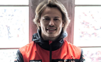 Portrait of a smiling man wearing a red and black jacket, standing indoors near a window.