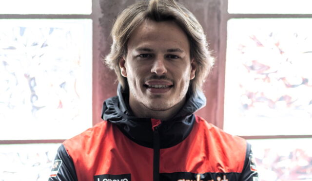 Portrait of a smiling man wearing a red and black jacket, standing indoors near a window.