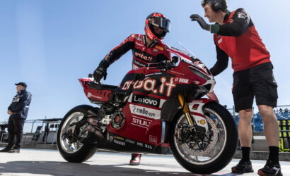 MotoGP rider in red sponsor livery sits on a red racing motorcycle in the pit lane as a crew member gestures beside the bike.