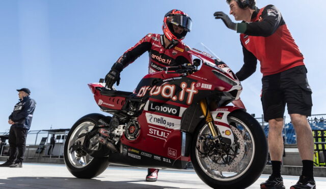 MotoGP rider in red sponsor livery sits on a red racing motorcycle in the pit lane as a crew member gestures beside the bike.