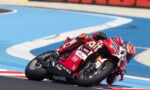 Red Ducati motorcycle racer leaning into a sharp turn on a blue race track, foreground close-up of the bike and rider's red suit with sponsor logos