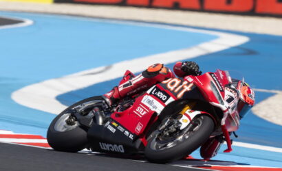 Red Ducati motorcycle racer leaning into a sharp turn on a blue race track, foreground close-up of the bike and rider's red suit with sponsor logos