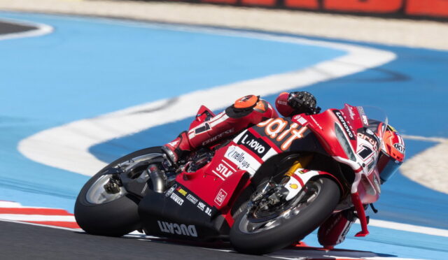 Red Ducati motorcycle racer leaning into a sharp turn on a blue race track, foreground close-up of the bike and rider's red suit with sponsor logos