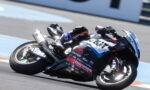 Motorcycle racer leaning into a turn on a racing track, full-suit and helmet with sponsor logos visible