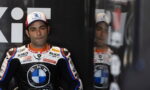 Moto racer in BMW/Rokit racing suit sits against black backdrop, wearing a white-blue-red cap, reflection visible in glass nearby.