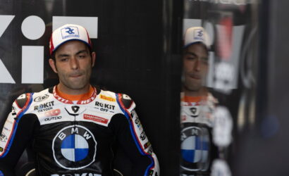 Moto race rider in BMW racing suit seated against a dark wall, reflection visible in glass beside him, wearing a white cap with blue logo.
