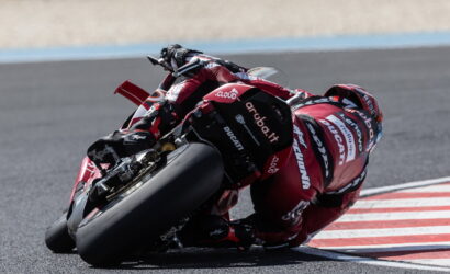 Motorcycle racer leaning into a sharp turn on a racetrack, wearing a red Ducati racing suit and bike visible in motion at speed.