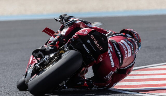 Motorcycle racer leaning into a sharp turn on a racetrack, wearing a red Ducati racing suit and bike visible in motion at speed.