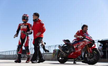Moto racer in red Honda racing suit and helmet walking beside a crew member on a sunny pit lane, with a red sport bike nearby on the right side of the frame.