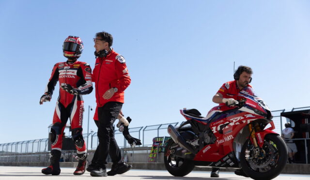 Moto racer in red Honda racing suit and helmet walking beside a crew member on a sunny pit lane, with a red sport bike nearby on the right side of the frame.