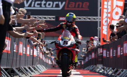 Motorcycle racer in red and black gear reaches out to fans as he crosses the finish line under a VictoryLane banner. - or - Rider on a red motorcycle with outstretched arms, greeted by cheering spectators along a barrier.