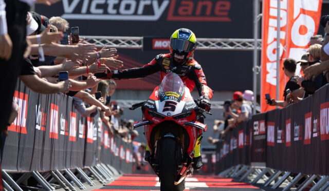 Motorcycle racer in red and black gear reaches out to fans as he crosses the finish line under a VictoryLane banner. - or - Rider on a red motorcycle with outstretched arms, greeted by cheering spectators along a barrier.