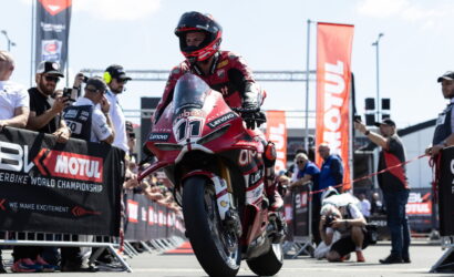 Motorcycle racer in red leathers on a racing bike, front wheel up, at a Moto 2/3 start with banners and spectators behind barriers