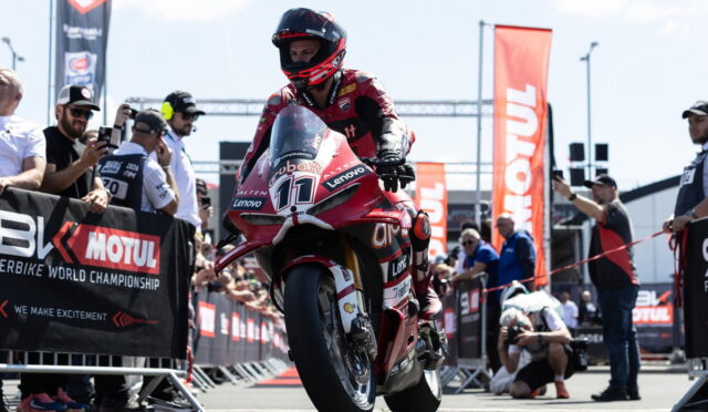 Motorcycle racer in red leathers on a racing bike, front wheel up, at a Moto 2/3 start with banners and spectators behind barriers