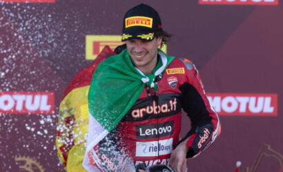 Racing driver on a podium spraying champagne, with an Italian flag draped over their shoulders and wearing a Pirelli cap and Ducati racing suit.