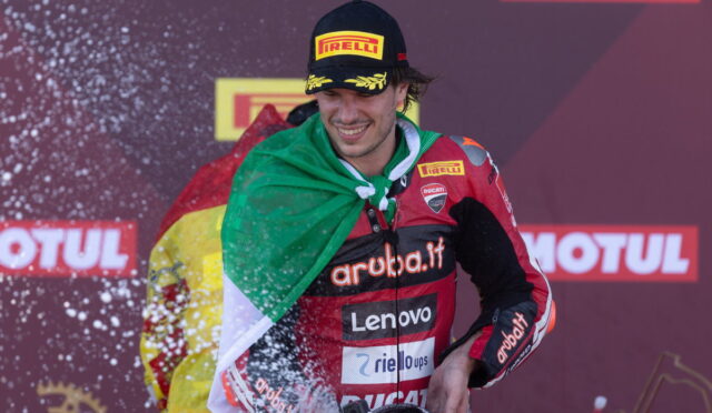 Racing driver on a podium spraying champagne, with an Italian flag draped over their shoulders and wearing a Pirelli cap and Ducati racing suit.