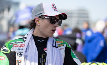 MotoGP rider in Kawasaki racing gear with white cap and sunglasses, looking to the side with a towel around the neck.