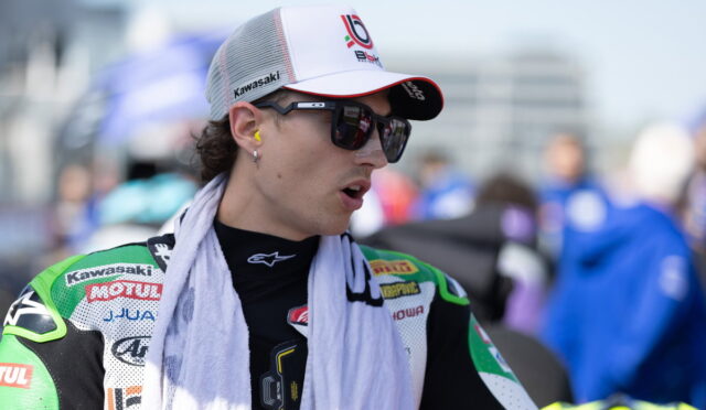 MotoGP rider in Kawasaki racing gear with white cap and sunglasses, looking to the side with a towel around the neck.