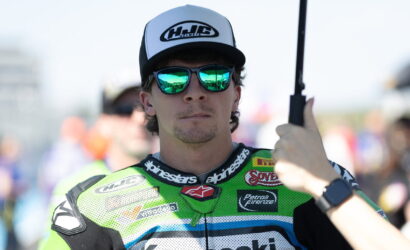 Portrait of a male motorcycle racer in a neon-green suit with a white cap and mirrored sunglasses, while someone holds a black umbrella over him.