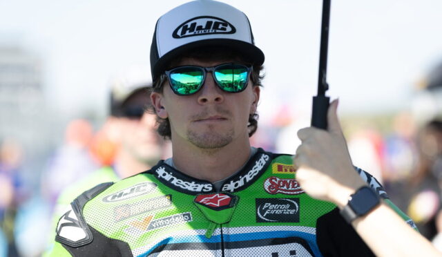Portrait of a male motorcycle racer in a neon-green suit with a white cap and mirrored sunglasses, while someone holds a black umbrella over him.
