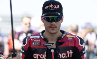 Male motorcycle racer in a red-black Aruba.it Racing suit and black cap with the Aruba.it logo, wearing mirrored sunglasses in a paddock.