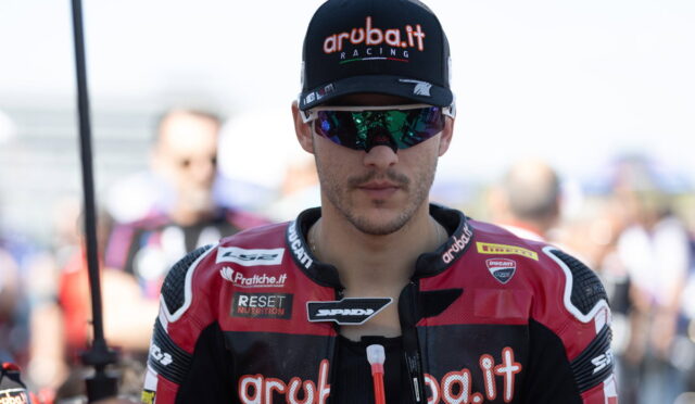 Male motorcycle racer wearing a black cap with aruba.it Racing and reflective sunglasses, facing the camera.
