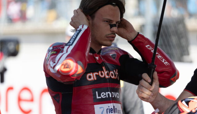 Moto racer in a red Aruba.it suit adjusts ear protection while a crew member holds an umbrella nearby.