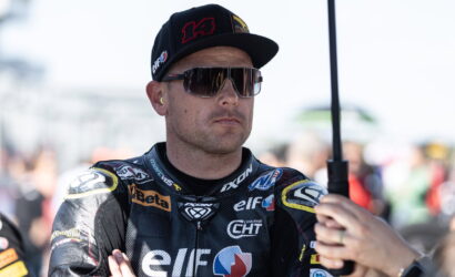 Racing driver in a black Elf racing suit with sponsor logos, wearing a cap and mirrored sunglasses, arms folded.
