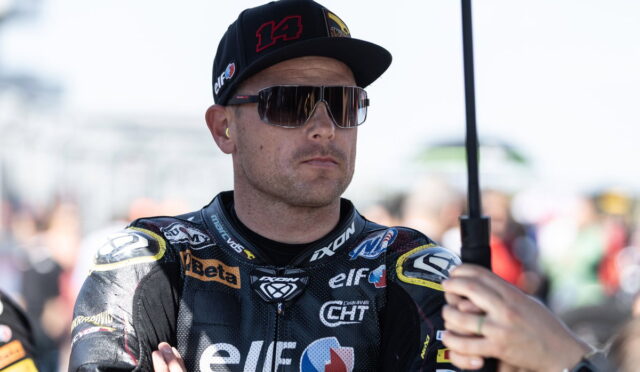 Racing driver in a black Elf racing suit with sponsor logos, wearing a cap and mirrored sunglasses, arms folded.
