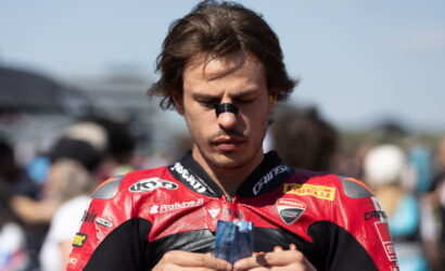 Motorcycle racer in a red Ducati suit concentrates on a blue item in his hands, crowd blurred in the background.