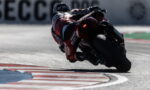 Motorcyclist in a red racing suit leans into a sharp corner on a racetrack at speed, tires gripping the asphalt.