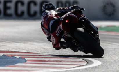 Motorcyclist in a red racing suit leans into a sharp corner on a racetrack at speed, tires gripping the asphalt.