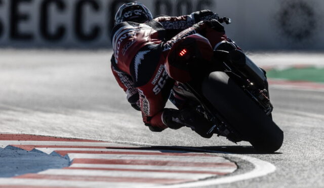 Motorcyclist in a red racing suit leans into a sharp corner on a racetrack at speed, tires gripping the asphalt.