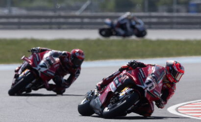 Racing motorcycles lean into a sharp turn on a race track, red bikes leading a group through a corner.