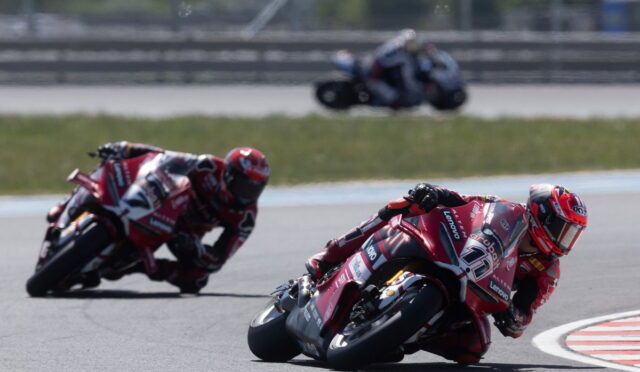 Racing motorcycles lean into a sharp turn on a race track, red bikes leading a group through a corner.