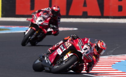 Two motorcyclists lean into a sharp turn on a race track, red bikes with racing livery, blue and red painted curbs in the foreground.