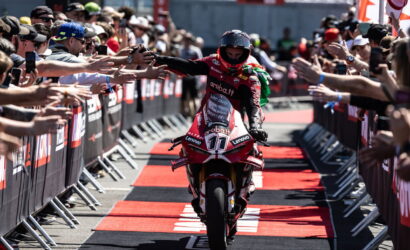 Motorcycle racer in red gear high-fives fans along a red-carpet finish line, crowds on both sides reaching out to celebrate a victory.